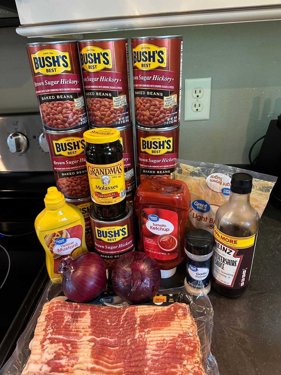 Ten cans of baked beans stacked with molasses, ketchup, mustard, brown sugar, garlic, red onions, Worcestershire sauce, and bacon